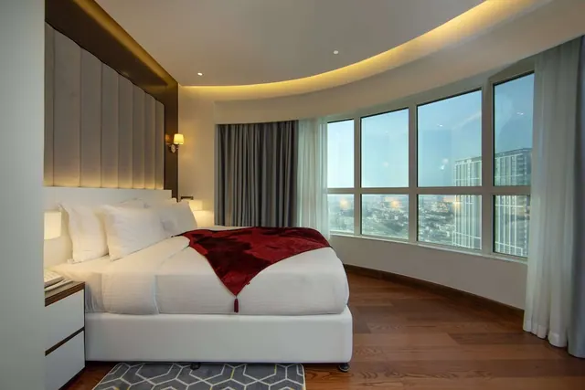 A king bed with white linens and a red throw sits in a bedroom with curved floor-to-ceiling windows overlooking city high-rises.