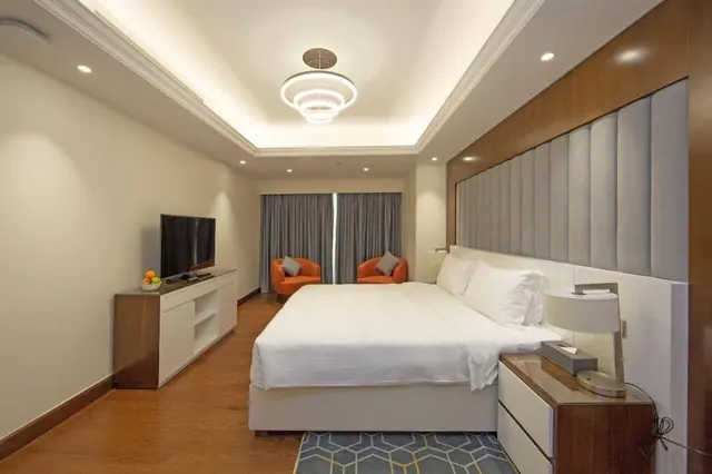 A bedroom with a king bed, two orange armchairs by the curtained window, a television on a white cabinet and a ring chandelier overhead.