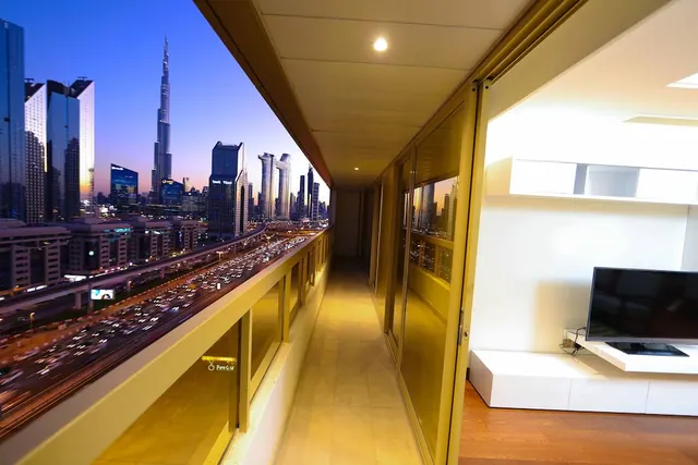 A long covered balcony with glass railings looks out over busy road traffic and the Burj Khalifa at dusk, with a wall-mounted television visible in the adjoining room.
