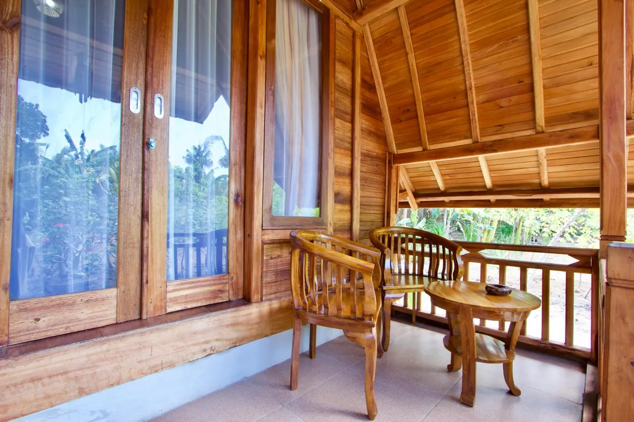 A covered wooden veranda with two wooden chairs and a small round table, with a railing overlooking green tropical trees.