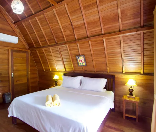 A bedroom with wood-panelled walls and a sloped wood ceiling, a double bed with white linen and two rolled towels, bedside lamps, and a wall-mounted air conditioner.