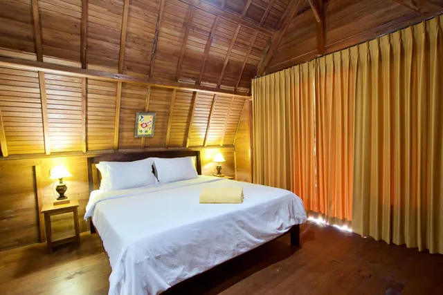 A bedroom with a timber ceiling and wooden walls holds a made bed with white linen and a folded yellow towel, two bedside lamps, and yellow curtains.