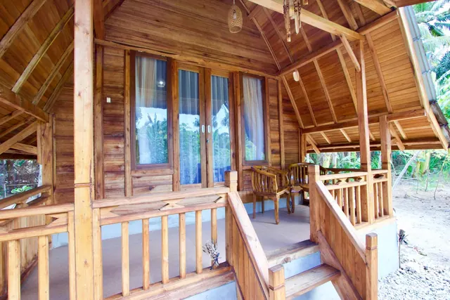A wooden veranda with railings, two wooden chairs and steps down the side, with glass doors and windows behind and palm trees visible outside.