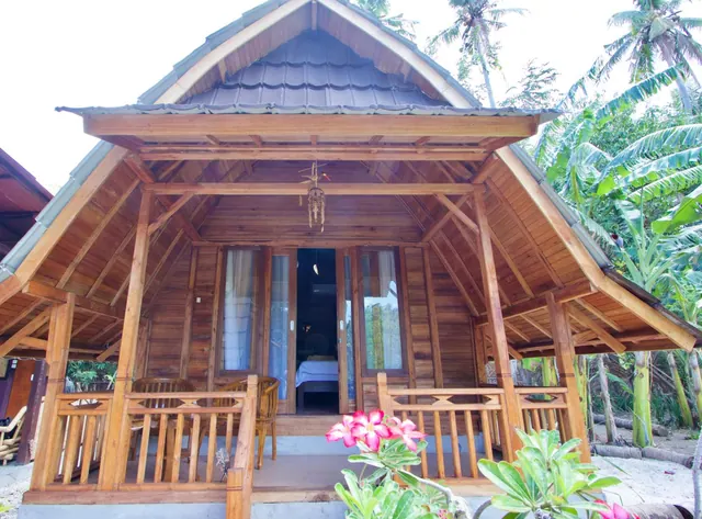 A traditional wooden bungalow with a steep dark-tiled roof and a front veranda with chairs stands among palm trees, with pink flowers in the foreground.