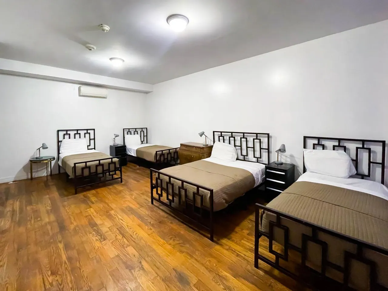 A bedroom with four single beds with dark geometric metal frames, bedside lamps, a wooden dresser, and a wall-mounted air conditioner under overhead ceiling lights.