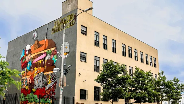 The exterior of a multi-storey building with a large mural on its side wall and a sign reading The New York Moore Hotel, with trees and a street lamp in front.