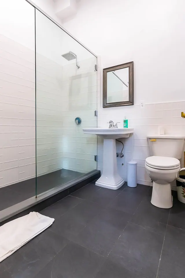A bathroom with a walk-in glass-enclosed shower, a pedestal sink beneath a framed mirror, and a toilet on a dark tiled floor.