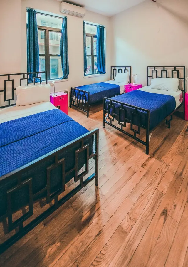 Three single beds with blue quilts and black metal frames sit in a room with wood floors, pink bedside tables, a wall air conditioner and two windows with blue curtains.