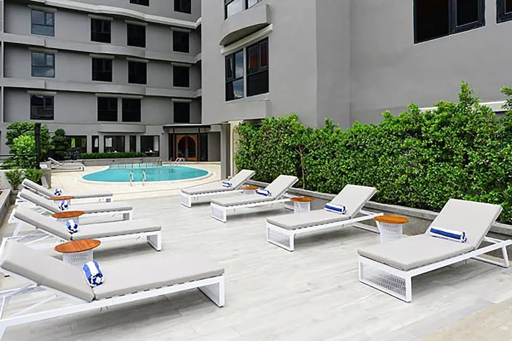 White sun loungers with grey cushions and blue-striped towels sit on a pale deck beside a small oval pool and a multi-storey building.
