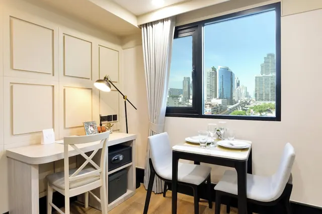 A hotel room with a desk, desk lamp, in-room safe and a dining table set for two beside a window overlooking city high-rises.