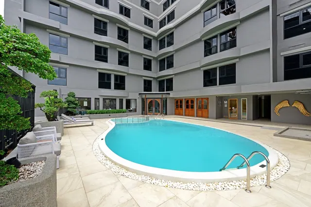 An outdoor kidney-shaped swimming pool with a white pebble border sits in a courtyard beside a grey multi-storey building, with sun loungers on the left.