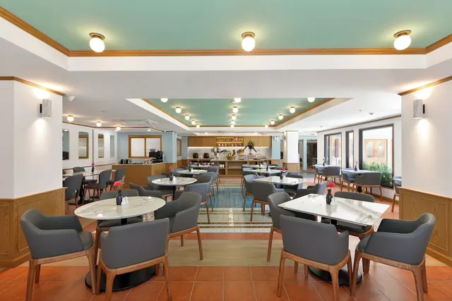 A dining room with round marble-top tables, grey upholstered chairs, a teal ceiling and a counter with coffee machines in the background.