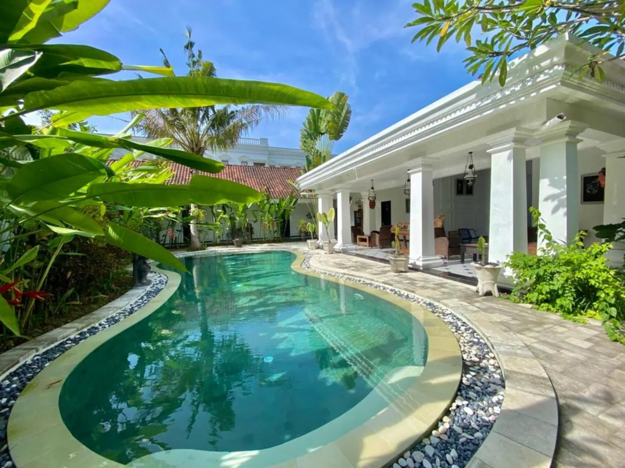 A kidney-shaped swimming pool with a pebble border sits beside a white colonnaded terrace with seating and tropical plants.