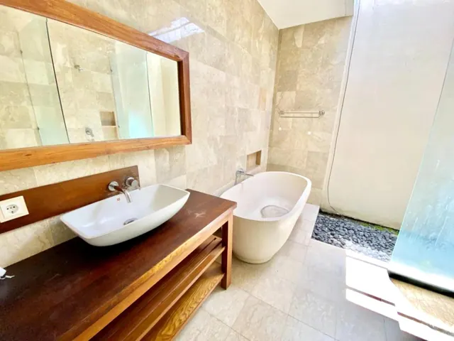 A bathroom with a wood-framed mirror, a vessel sink on a wooden vanity, a freestanding white bathtub and an open shower area with pebble flooring.