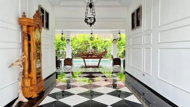 A tiled entrance hall with a black and white checkered marble floor, a carved wooden grandfather clock, and an open view through to a courtyard with plants, a wooden table and a pool.