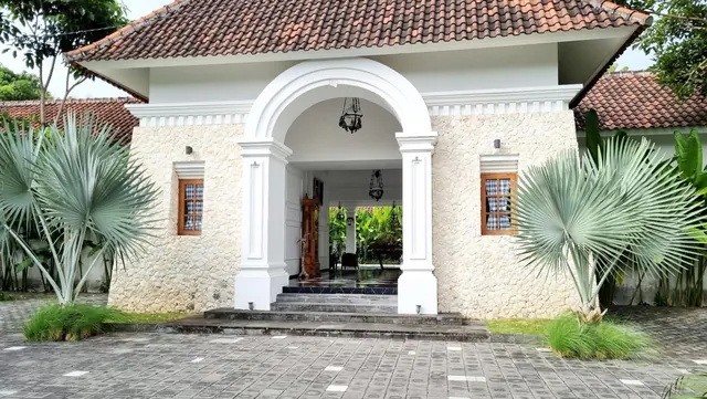A white arched entrance with columns sits in a stone-walled building with a terracotta tiled roof, flanked by fan palms on a paved forecourt.