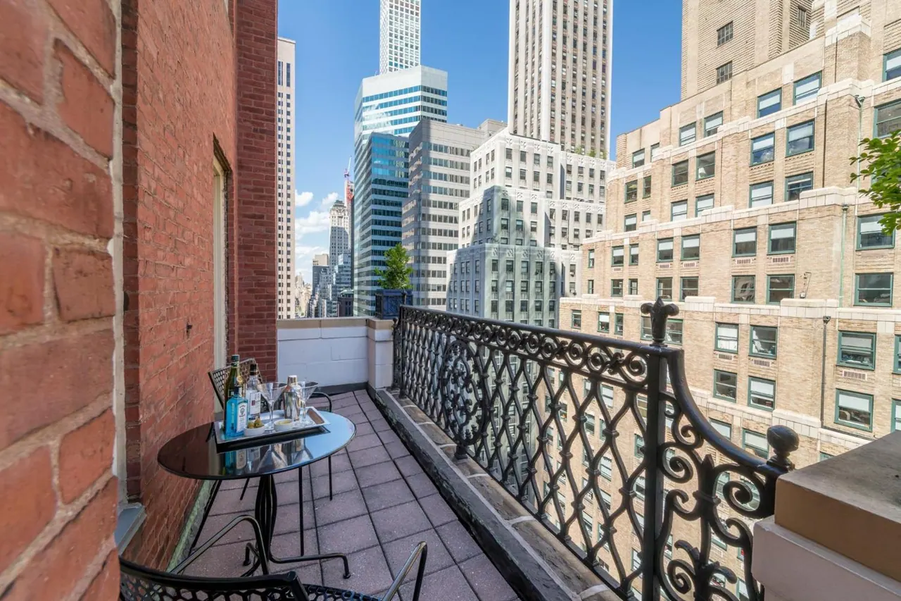 A narrow private balcony with a small round glass table, two chairs and a wrought iron railing overlooks high-rise buildings and a city skyline.