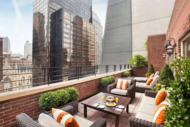 A rooftop terrace with wicker seating, orange striped cushions and a set dining table, overlooking skyscrapers and a Gothic cathedral.
