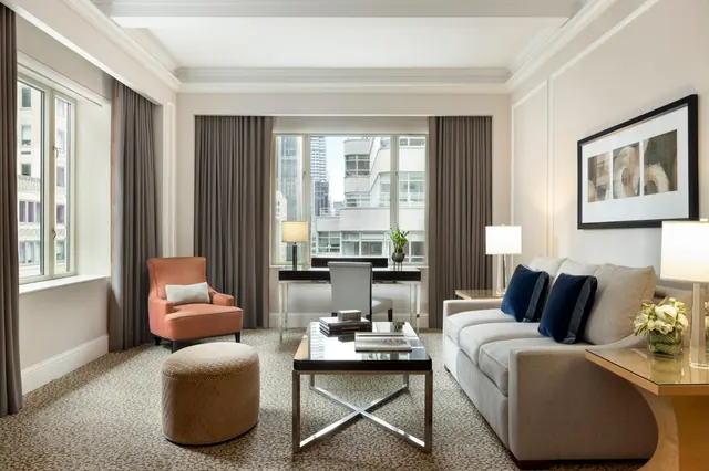 A living area in a hotel suite with a grey sofa, blue cushions, a coral armchair, an ottoman and a coffee table, with a desk by a window looking out onto city buildings.