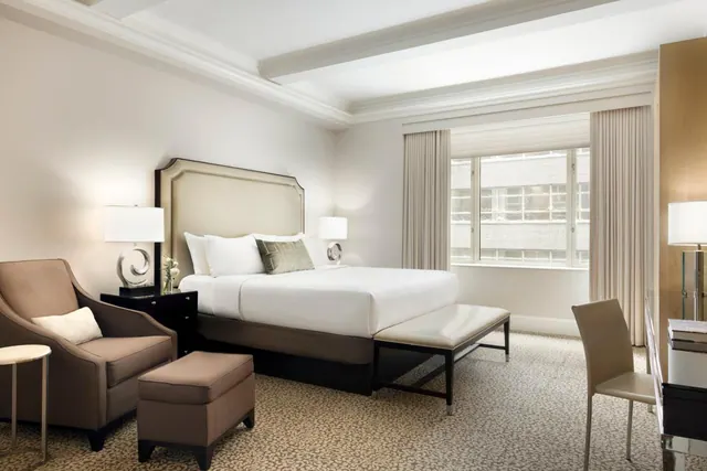 A bedroom with a king bed, an upholstered armchair with an ottoman, a desk and chair, and a window facing a building across the street.