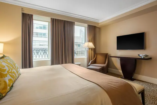 A king bed with a yellow patterned pillow faces a wall-mounted television, an armchair and windows overlooking city buildings.