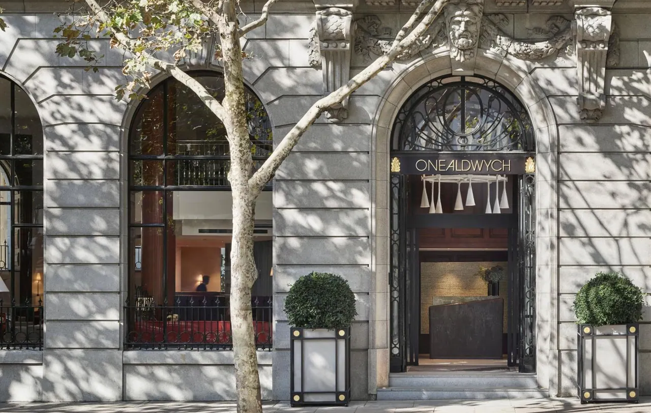 The arched stone entrance of the One Aldwych hotel has its name on a sign above the doors, with two topiary planters on the pavement and a tree in front.