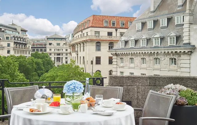A breakfast table with white linen and a blue flower arrangement sits on a balcony overlooking historic city buildings and trees.