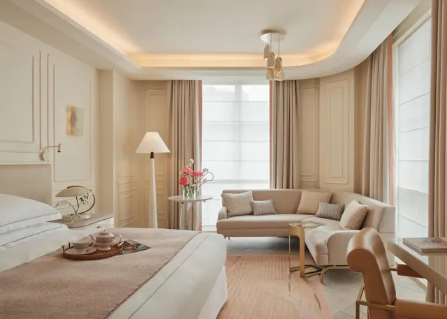 A bedroom with a made king bed and a tea tray on the bed, a curved beige sofa with cushions, a floor lamp, and a desk with a leather chair by the window.