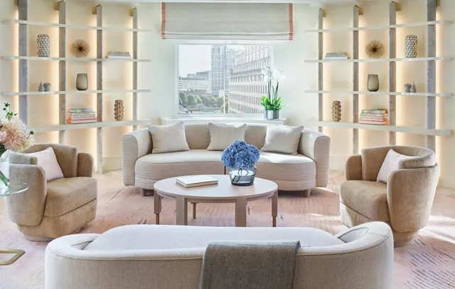 A living room with a curved sofa, two armchairs, a round coffee table with blue flowers, lit open shelving, and a window showing city buildings.