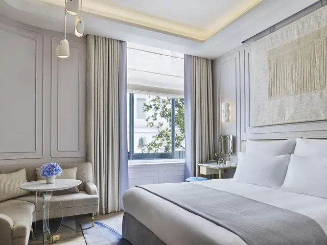 A bedroom with a made king bed, a grey upholstered sofa beside a marble side table holding blue flowers, and a window with a tree visible outside.