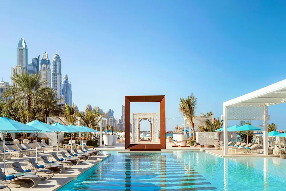 A large pool with teal umbrellas and sun loungers beside a white pergola, with a row of high-rise towers and palm trees behind.