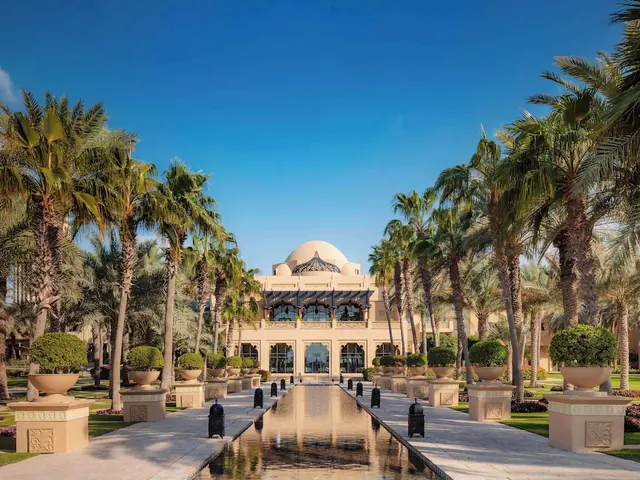A domed hotel building stands at the end of a paved walkway with a long narrow reflecting pool, lined by palm trees and topiary in stone urns.