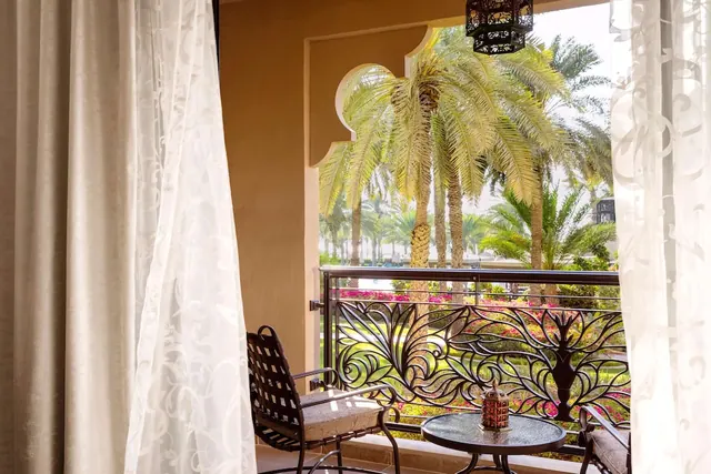 A private balcony with two metal chairs and a small glass table with a lantern, framed by sheer curtains and overlooking palm trees and pink flowering shrubs.