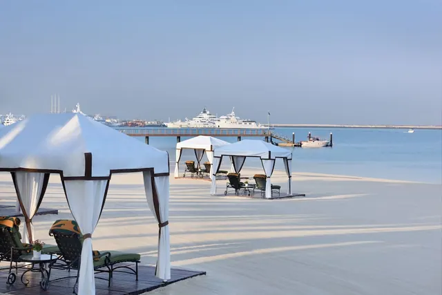 White canopy cabanas and cushioned lounge chairs stand on a pale sandy beach facing calm sea, with moored yachts and a jetty in the distance.