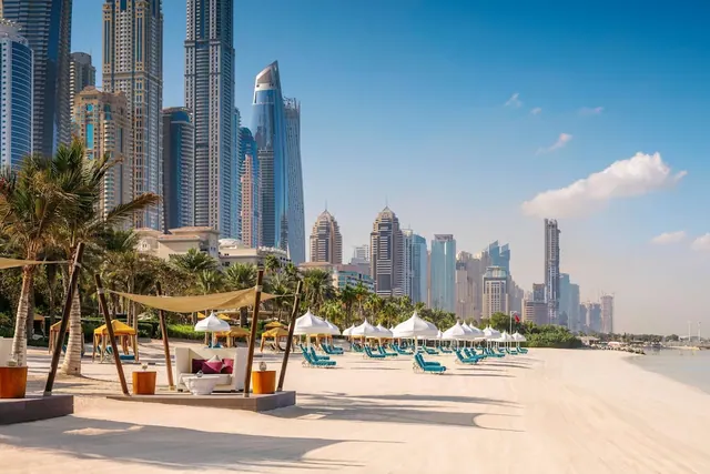 A sandy beach with white umbrellas, teal sun loungers and a row of high-rise towers behind it under a clear blue sky.