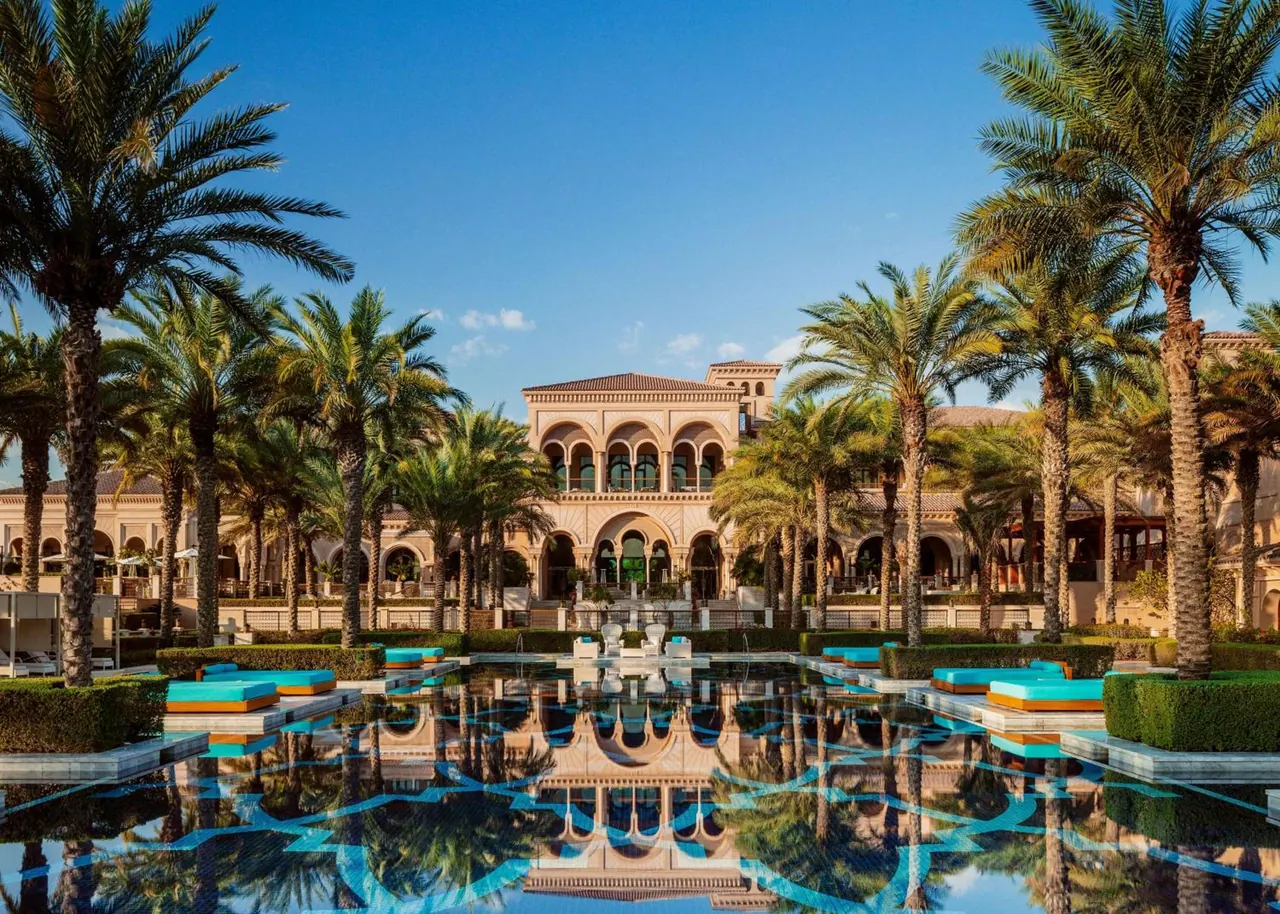 A long reflecting pool lined with turquoise sun loungers and palm trees, with an arched Moorish-style building behind it under a clear blue sky.