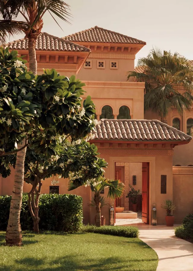A terracotta-coloured building with tiled roofs stands behind palm trees, flowering frangipani, a lawn and a paved pathway.