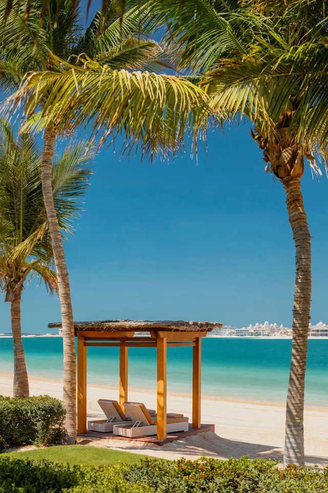 A thatched wooden cabana with two sun loungers stands on a sandy beach beside turquoise sea, framed by palm trees, with a distant resort building across the water.