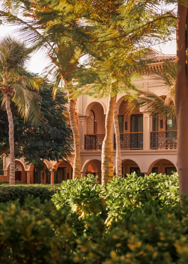 Exterior of a two-storey building with arched colonnades and ornate balcony railings, partly framed by palm trees and shrubs in the foreground.