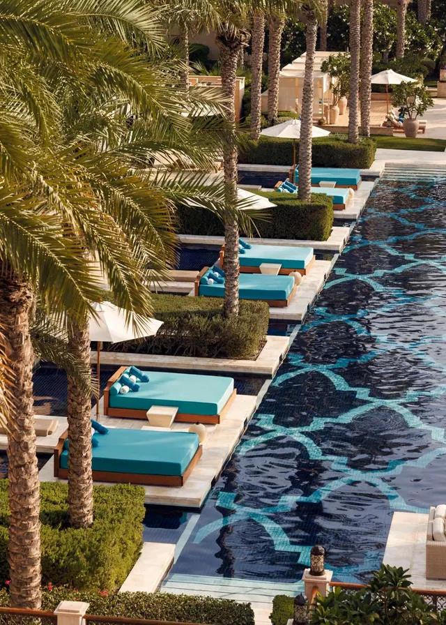 Turquoise-cushioned daybeds and white parasols line a long pool with a blue patterned tiled bottom, with palm trees along the edge.