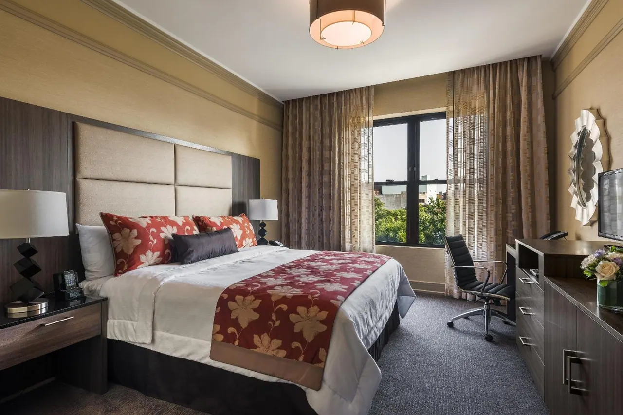 A bedroom with a king bed dressed in red floral throws, a black office chair by a window looking out at buildings and trees, and a wall-mounted television beside a dresser.