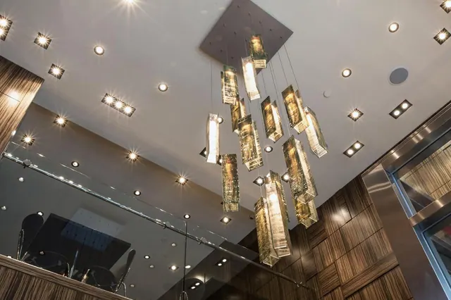 A cascading glass chandelier hangs beneath a ceiling with recessed downlights, beside wood-veneer walls, a glass railing and a lift.
