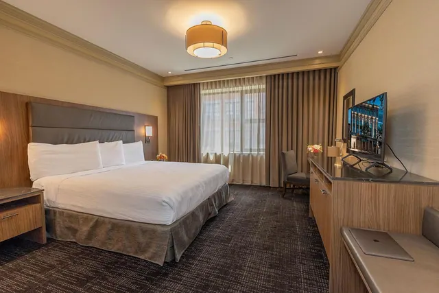 A king bed with white linens sits beside a window with sheer curtains, opposite a wooden desk with a wall-mounted television and a chair.
