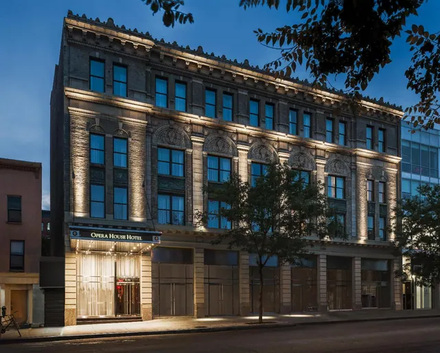 The ornate stone facade of the Opera House Hotel at dusk, with its illuminated entrance canopy and sign at street level.