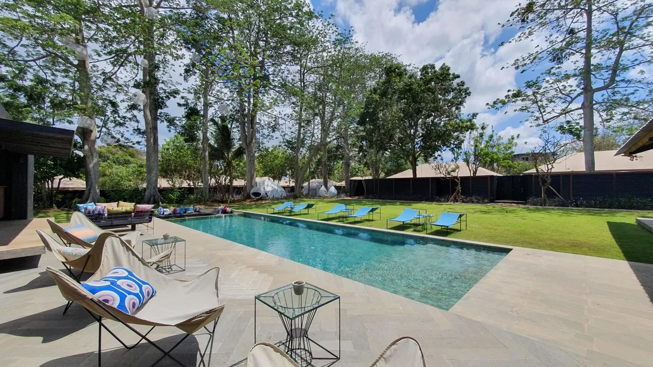A long rectangular swimming pool with blue sun loungers on a lawn, surrounded by trees, with outdoor armchairs and side tables on a stone deck.