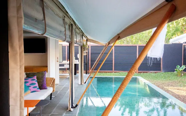 A covered terrace with a sofa and colorful cushions sits beside a small swimming pool, with a grass garden and slatted fence behind.
