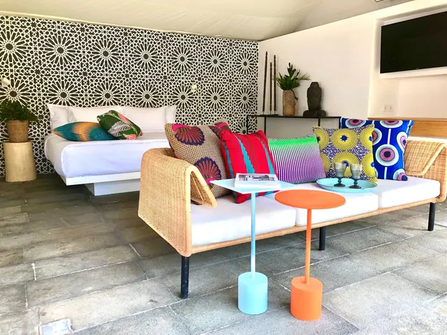 A wicker sofa with colourful patterned cushions and two side tables sits in front of a bed against a black and white patterned wall.