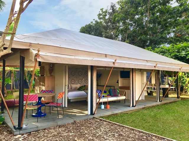 A canvas-roofed guest unit with an open front terrace shows a bed, a sofa with cushions, and outdoor dining chairs on a paved floor beside a lawn.