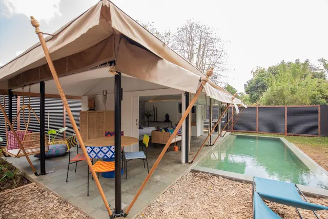 A tent-style villa with a canvas roof, an outdoor dining table and chairs, and a long rectangular pool with blue sun loungers in the foreground.