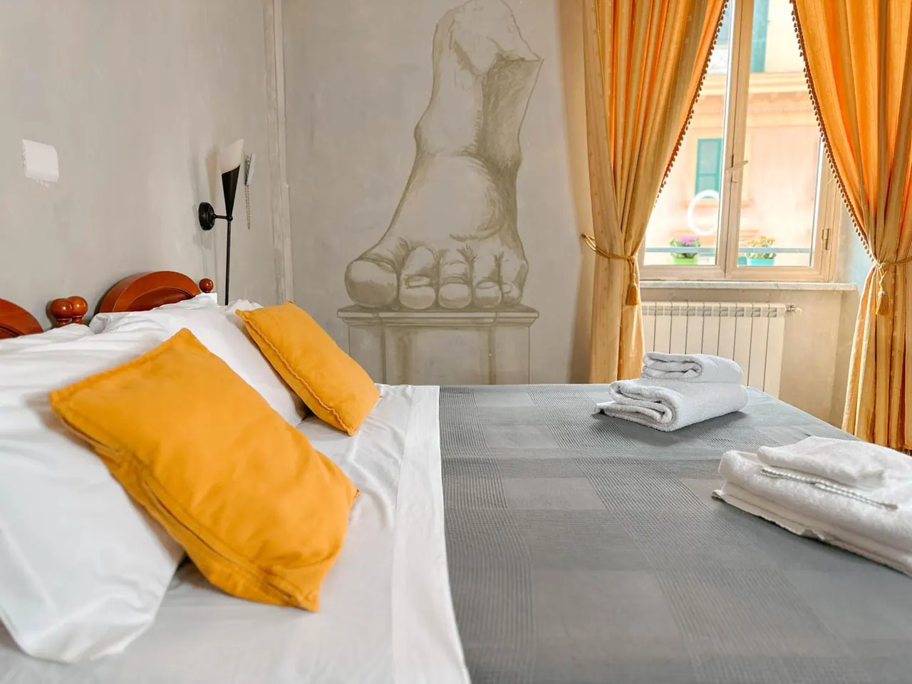 A bedroom with a double bed, orange cushions and folded white towels on a grey bedspread, a wall mural of a foot, and an orange-curtained window looking onto a building facade.